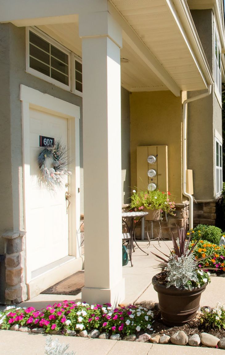 Entrance of an apartment with potted plants, flowers, and a pathway leading to a sunny, tree-filled complex.