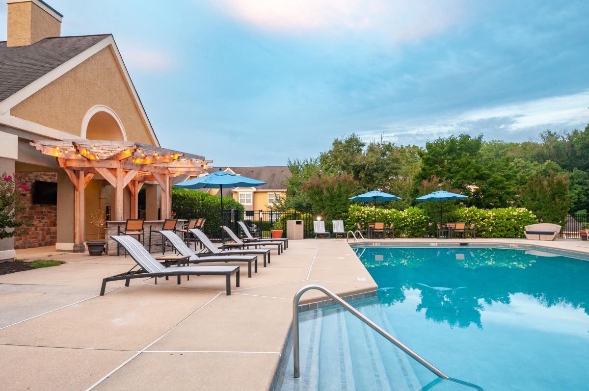 Outdoor pool with lounge chairs, shaded patio, and blue umbrellas surrounded by greenery at sunset.