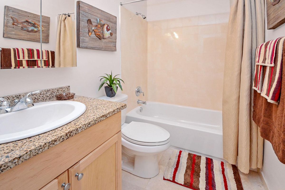 Small bathroom with granite countertop, bathtub, striped towels and rug, fish wall art, and a potted plant.