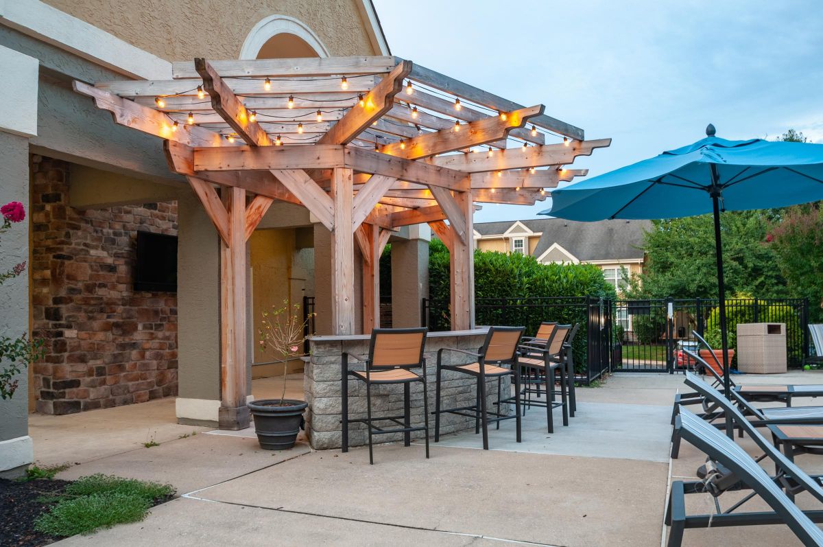Wooden pergola with string lights over a bar and chairs beside a pool area with a blue umbrella.