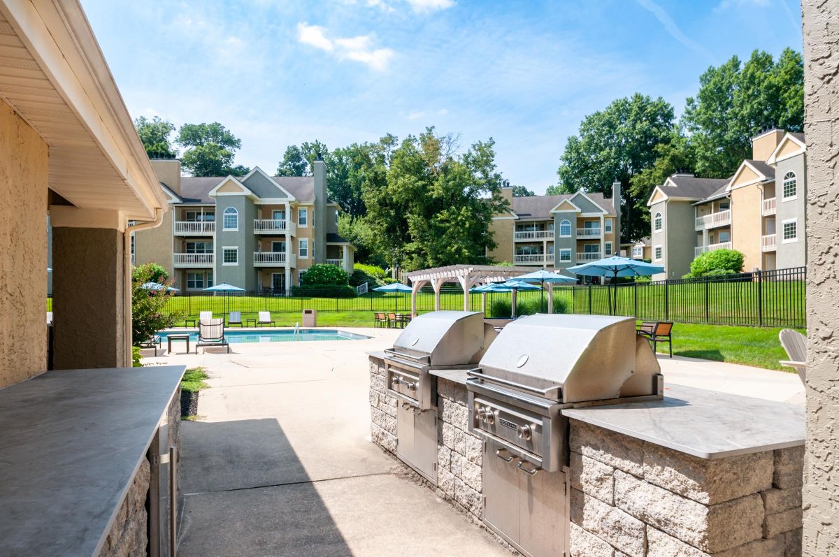 Outdoor grilling area near a pool with lounge chairs, umbrellas, and apartment buildings in the background.