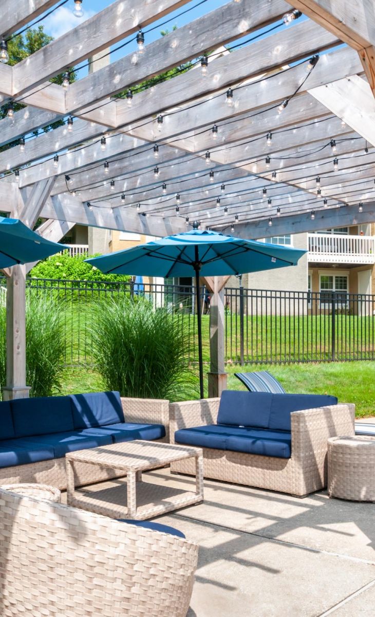 Outdoor patio with wicker seating, blue cushions, blue umbrellas, and a wooden pergola beside a pool area.