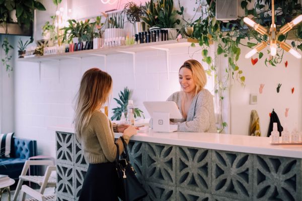 Two women interact at a modern, plant-filled salon reception desk with shelves of products behind them.
