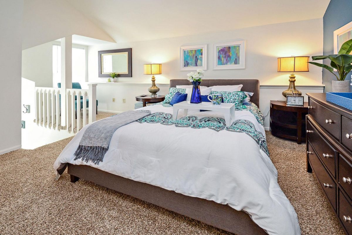 Bright bedroom with a neatly made bed, bedside tables, lamps, and modern decor on carpeted flooring.