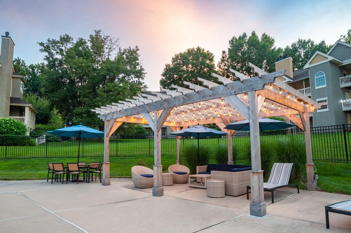 Wood pergola with string lights over outdoor seating and tables, near green lawn and apartments at sunset.